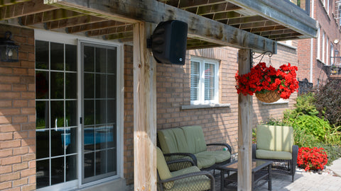Outdoor patio area with a wooden pergola, chairs, and potted plants and a PSB CS100 outdoor speaker mounted on the post